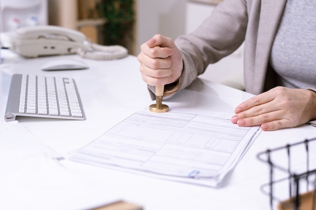 Foto handen van jonge vrouwelijke beambte die door bureau zit en zegel op financieel document zet alvorens het naar cliënt te verzenden