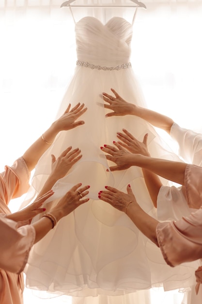 Handen van onherkenbare vrouwen met armbanden, ringen en manicure die een verbazingwekkende witte trouwjurk aanraken die aan een hanger tegen het raam in de zonovergoten kamer hangt. Bijeenkomst en voorbereiding van de ochtend van de bruid