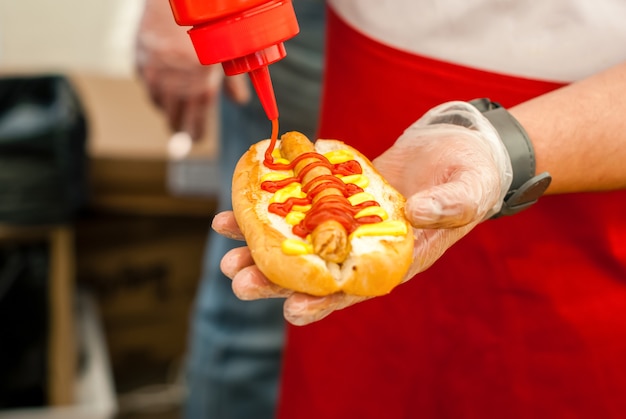 Handen van straatchef die ketchup op hotdog van dichtbij toevoegt