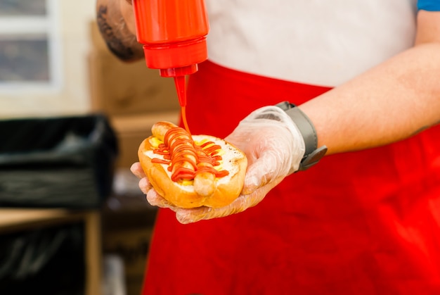 Handen van straatchef die ketchup op hotdog van dichtbij toevoegt