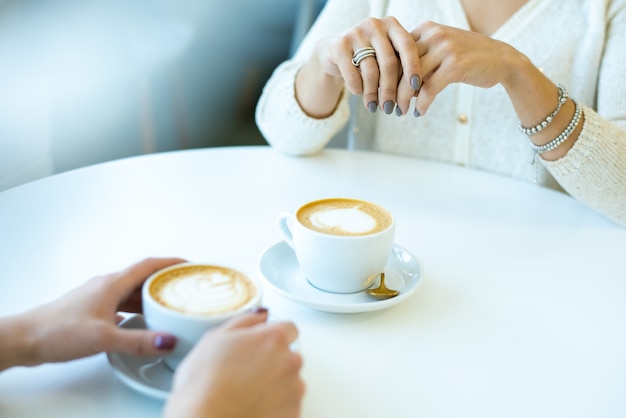 Handen van twee jonge vriendelijke vrouwen in vrijetijdskleding aan tafel in café zitten terwijl het hebben van cappuccino tijdens gesprek tijdens pauze