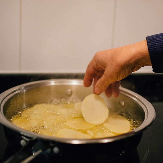 Handen van volwassen vrouw die gesneden aardappelen toevoegt in een koekenpan met hete olie in de keuken