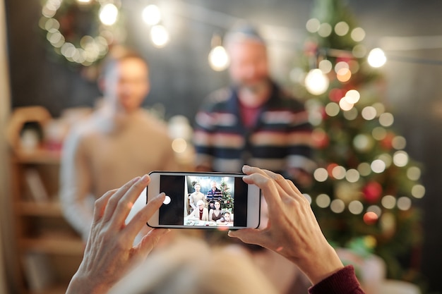 Handen van vrouwelijke bedrijf smartphone met foto van aanhankelijk en gelukkige familie op het scherm tijdens het nemen van foto door feestelijke tafel