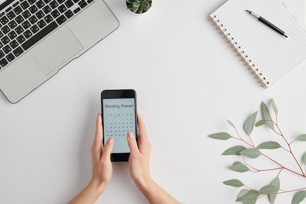 Handen van vrouwelijke werknemer met smartphone met maandelijkse planner op scherm over wit bureau tijdens het werk