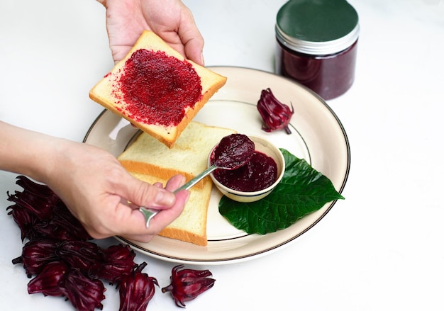 Handen verspreiden hibiscusjam op een toast tegen pot met jam en hibiscusbloemen
