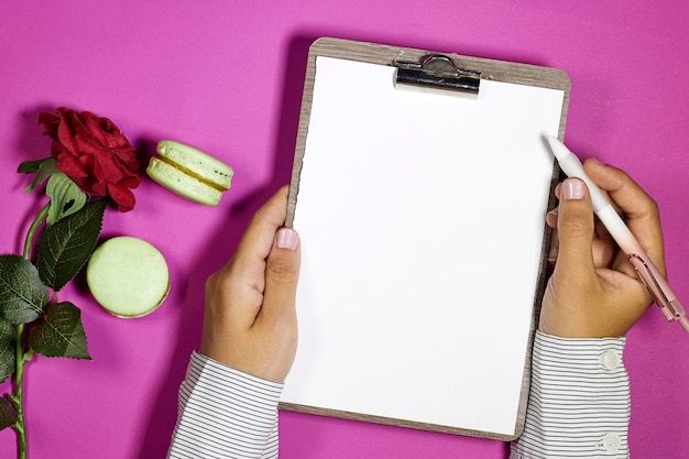 handen vrouw met een klassiek houten papierklembord met rode roze bloem en macaron op paarse backgr