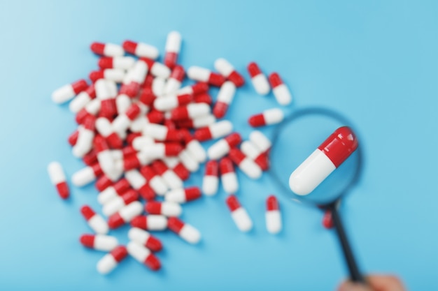 A handful of Red and white pill capsules are examined with a magnifying glass on a blue background. Close-up. Top view