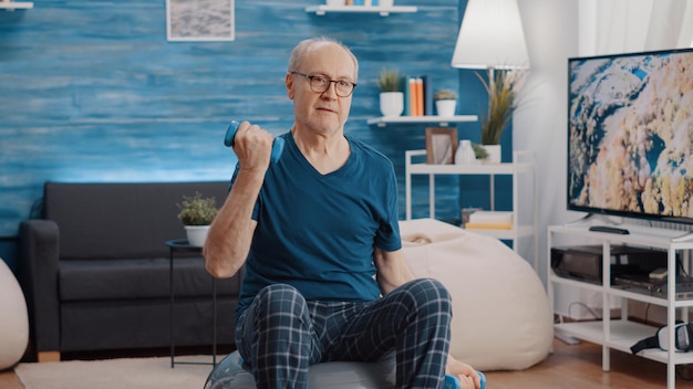 Handheld shot of old man lifting weights and training while sitting on fitness toning ball at home. Senior person using dumbbells to train and do physical exercise, stretching arms muscles