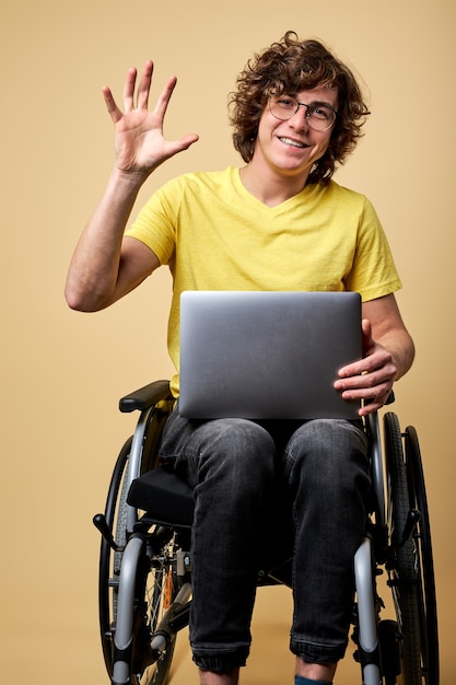 Handicapped male on wheelchair say hello sitting on wheelchair with modern laptop, working online , smiling.