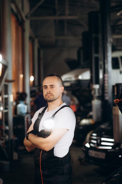 Handige auto-mechanicus poseert in een auto-service hij draagt een spijkerbroek hemd en veiligheidsbril zijn armen gekruist specialist kijkt naar een camera en glimlacht