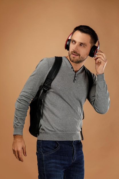 Foto handige jonge man in koptelefoon met rugzak luistert naar muziek geïsoleerd op beige achtergrond