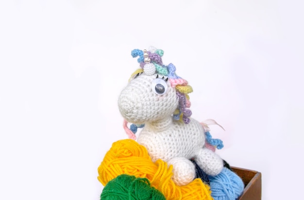 Photo handmade crocheted unicorn toy and colorul yarn in a wooden box on white background.