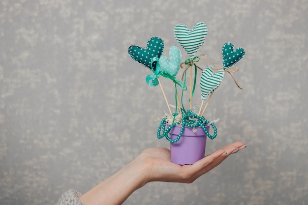 Handmade fabric hearts on sticks in a pot. The girl holds a handicraft in hand on a gray background.