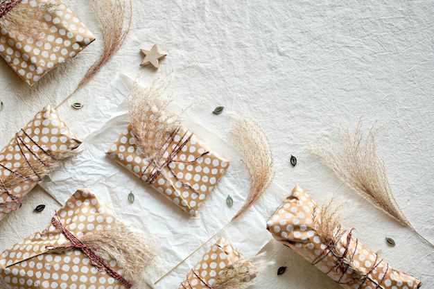 Handmade gifts wrapped in beige craft paper with dry wild grass\
tied up with cotton cord geometric flat lay on off white textile\
with natural decorations