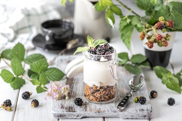 Handmade granola with white natural yogurt with blackberries in a glass transparent jar, flowers and leaves on a white wooden background.