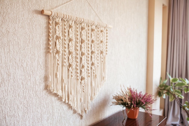 Handmade macrame wall decoration with wooden stick hanging on a white wall Female hobby ECO friendly modern knitting natural decoration in interior Copy space