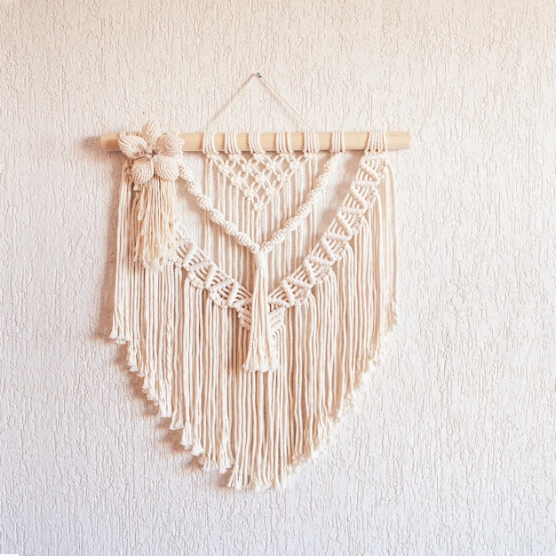 Handmade macrame wall decoration with wooden stick hanging on a white wall Macrame braiding and cotton threads Female hobby ECO friendly modern knitting natural decoration in interior Copy space