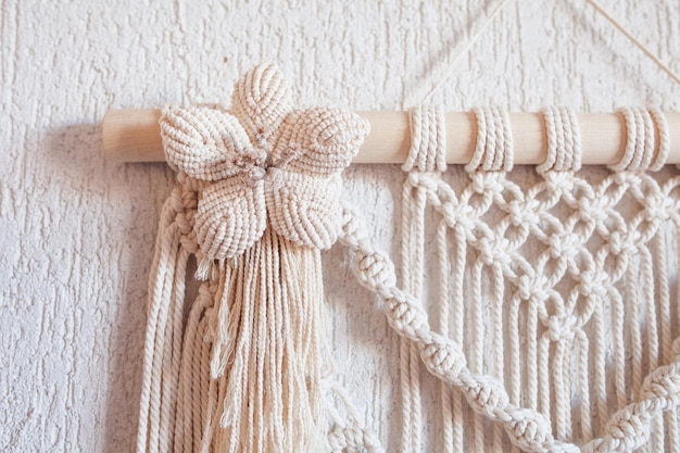 Handmade macrame wall decoration with wooden stick hanging on a\
white wall macrame braiding and cotton threads female hobby eco\
friendly modern knitting natural decoration in interior