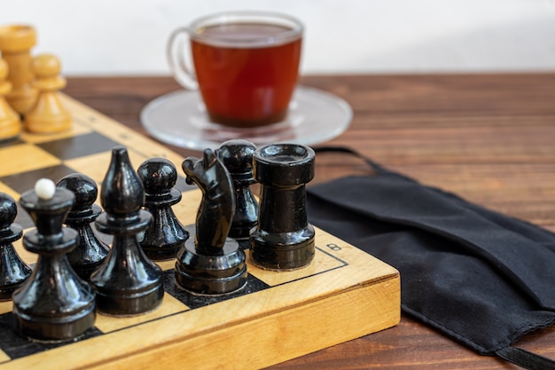 Handmade old chess on a wooden board. Occupation on vacation and quarantine. Nearby is a cup of coffee and a medical mask. The concept of isolation.