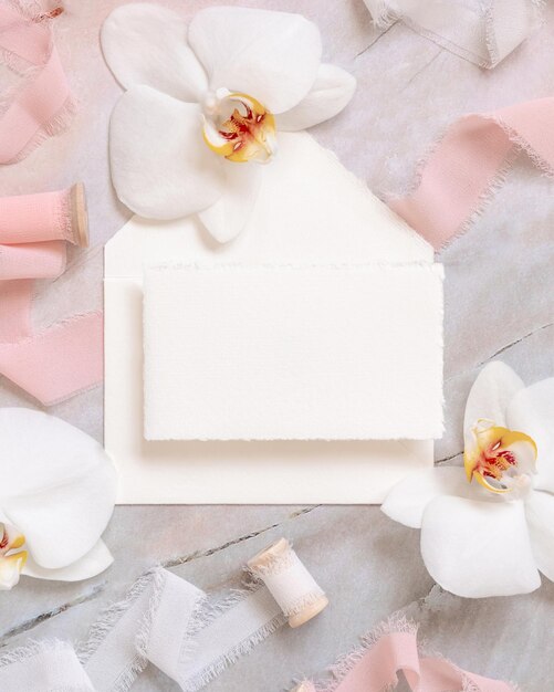 Handmade paper card and envelope near white orchid flowers and silk ribbons on marble mockup