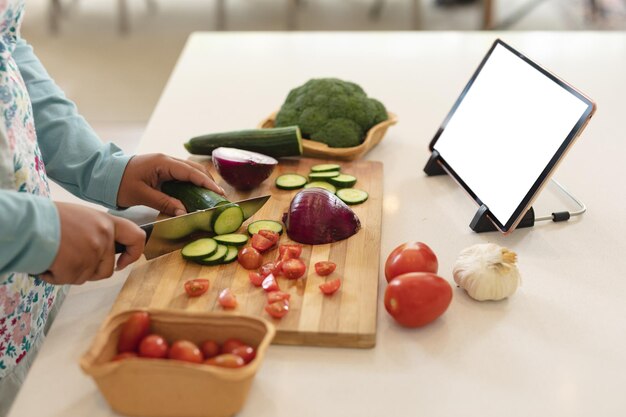 Photo hands of african american woman using tablet with copy space and cutting vegetables in kitchen