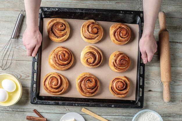 Hands are holding a baking tray with ready made cinnamon rolls buns over a wooden table with ingredients Concept of delicious homemade pastries and a cozy atmosphere Top view