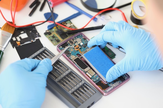 Hands in blue gloves with tweezers are repairing the battery
from a smartphone, close-up. mobile phone repair service