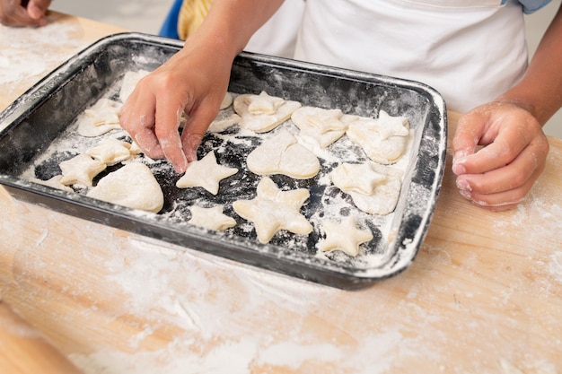 Mani del ragazzo che tagliano i biscotti dalla pasta arrotolata durante la cottura della pasticceria