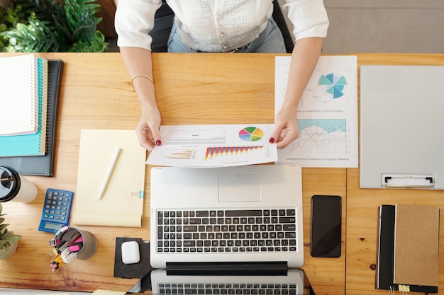 Hands of businesswoman reading and analyzing document with bar chart and circle diagram when working at table with opened laptop