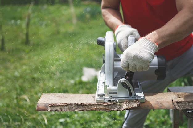 Hands of a carpenter's man in gloves sawing with an electric disk saw. Red T-shirt, gray pants, against the background of green grass and trees. Handwork, construction of houses, tools.