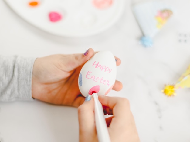 The hands of a caucasian girl in a gray turtleneck write with a pink marker on the egg the inscription Happy Easter