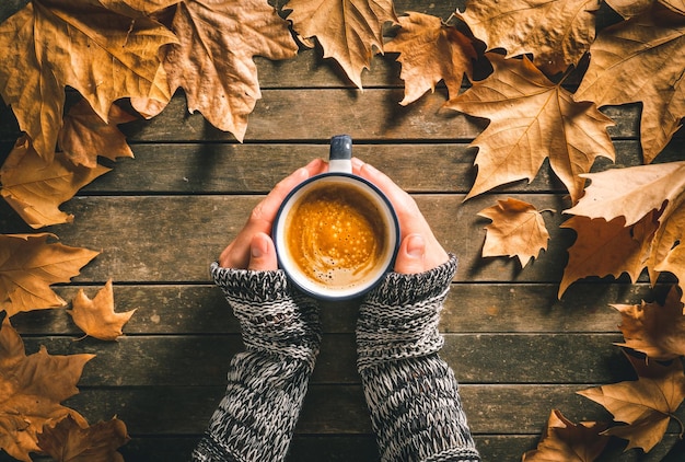 Hands of a caucasian male holding a hot coffee cup on an aged wooden table with autumn leaves around