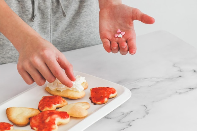 Hands of a caucasian teenage girl laying coffee cups heart sprinkles on not perfect freshly baked heart cookies