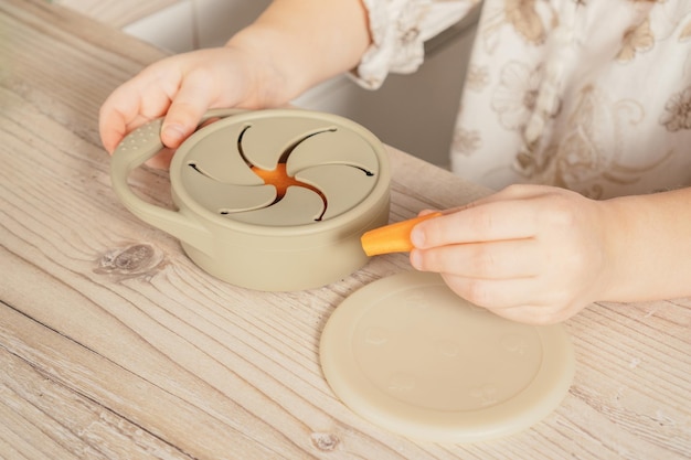 Hands of child take piece of carrot out from pastel gray silicone snack cup at wooden table Baby accessories tableware