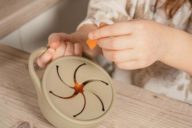 Hands of child take piece of carrot out from pastel gray silicone snack cup at wooden table Baby accessories tableware
