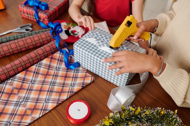 Photo hands of children using glue gun when wrapping presents for christmas in checkered paper