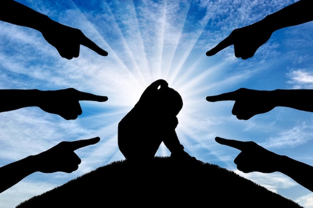 Hands of classmates point their fingers at the sad and lonely child girl sitting on the hill. Conceptual image of bullying children