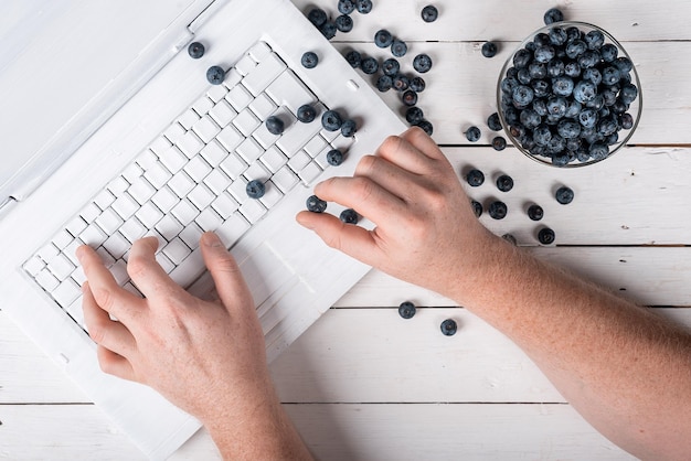 나무 흰색 테이블에 있는 컴퓨터와 블루베리에 손을 대고 건강한 스낵 개념