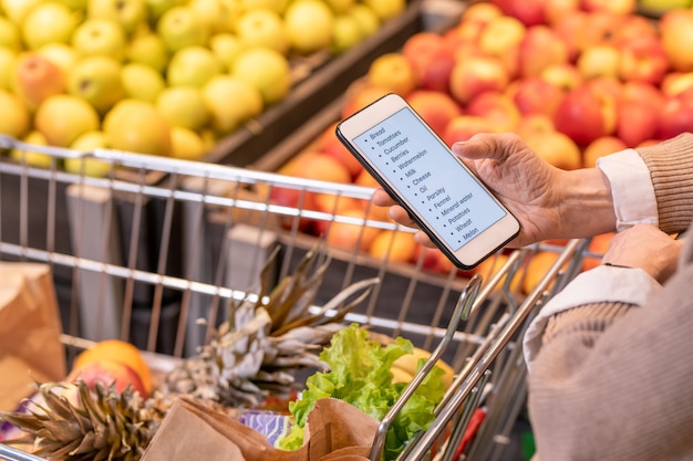 Hands of contemporary aged woman looking through shopping list in smartphone over cart with fresh fruit and vegetables in supermarket