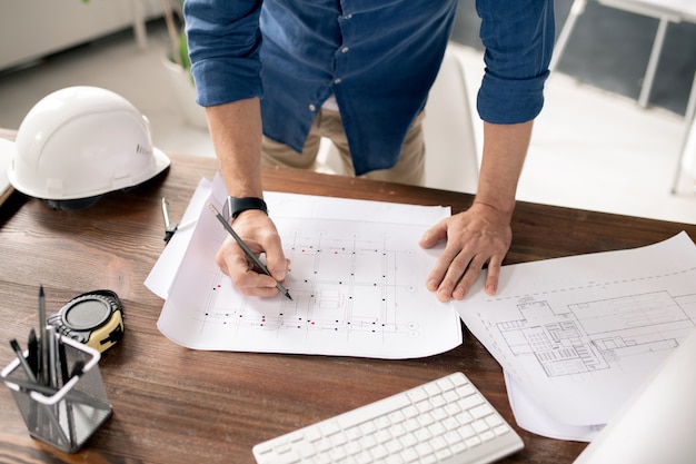 Hands of contemporary architect with pencil or crayon drawing sketch of building construction while standing by table