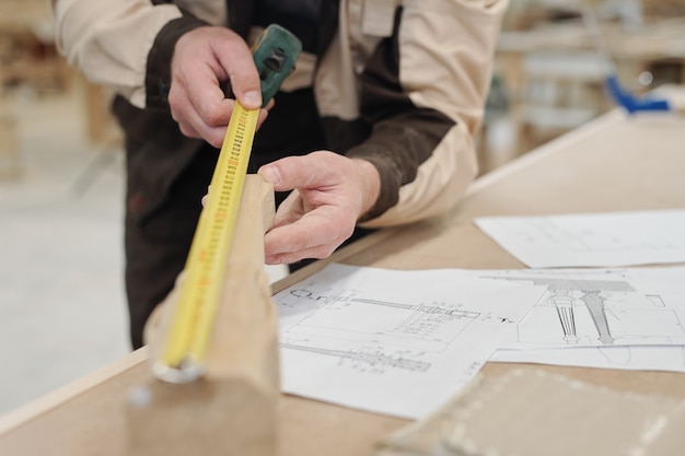 Hands of contemporary worker of factory holding wooden workpiece and measuring its length by workplace with sketches of this detail