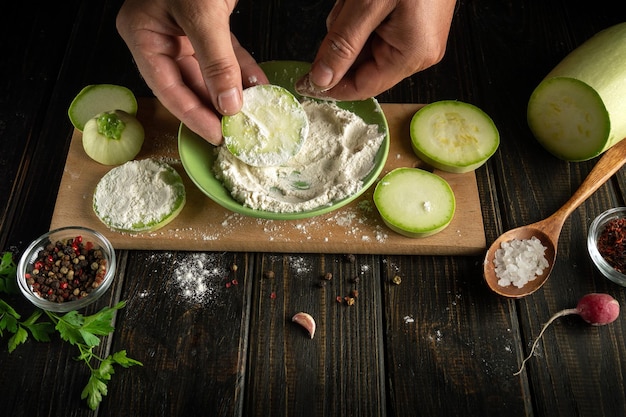 The hands of the cook add flour to the zucchini before fire with spices and vegetables Working environment on the kitchen table with vegetable marrow