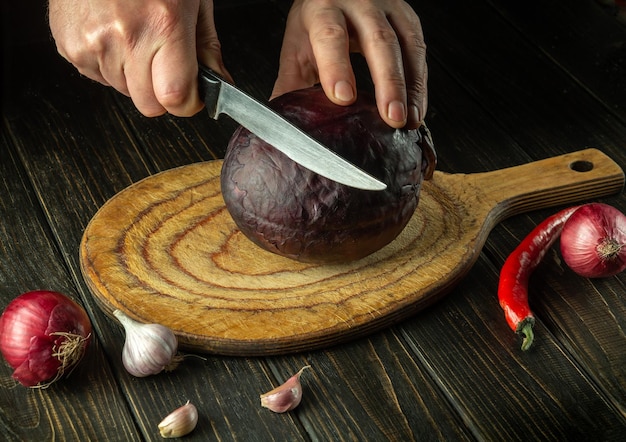 The hands of the cook are cutting red cabbage with a knife on a kitchen cutting board