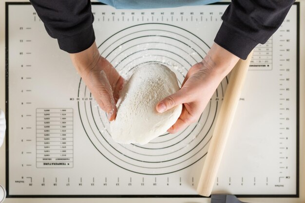 The hands of a cook in a blue apron hold wheat dough over a silicone baking mat top view selective focus