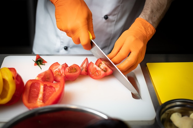 Mani di un cuoco in guanti, fette di pomodori su un tagliere bianco, in cucina