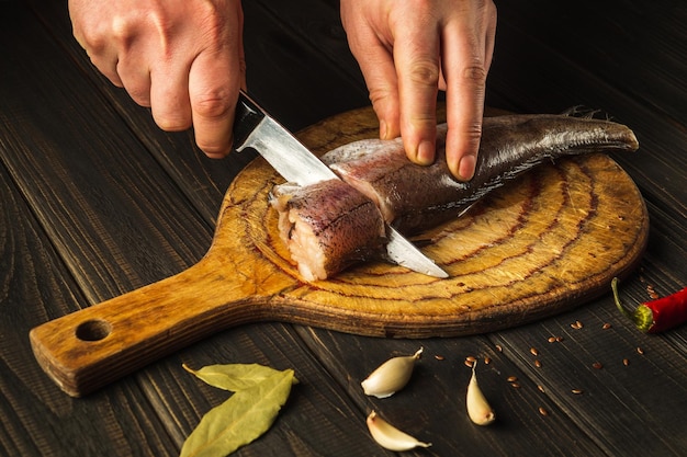 The hands of the cook with a knife cut fresh Merluccius fish on a wooden cutting board