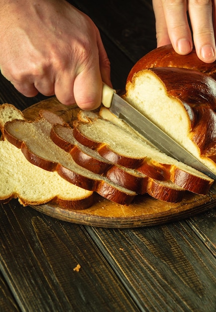 Руки повара с ножом режут свежий пшеничный хлеб или калач на кухонной доске для резки Здоровая еда или концепция приготовления пищи