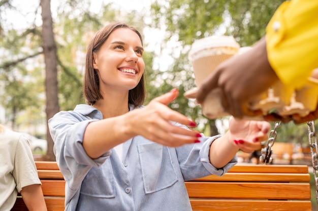 Hands of courier in yellow jacket passing two drinks from restarant or cafe to young smiling brunette woman sitting on wooden bench in park