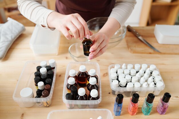 Hands of craftswoman opening small bottle with chosen aromatic essence over plastic containers on wooden table