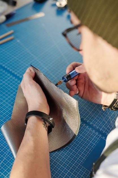 青いテーブルのそばに座って、クレヨンで灰色の革片のエッジを処理するブレスレットの創造的な革細工人の手
